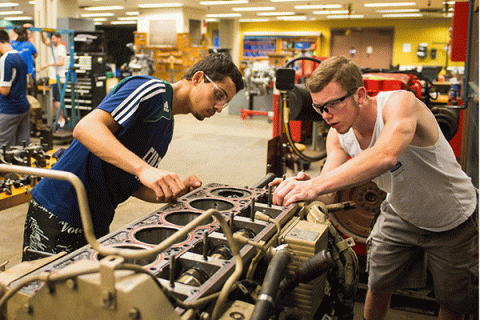 What looks like a a radiator or air compressor – grey with round and square uniform openings in the top – and with assorted green and black valves, hoses, and attachments – sits in a large industrial workspace with yellow walls. Two male students wearing safety goggles lean over it with their hands on it, inspecting something on the far end. The student on the left has short black hair and some facial hair and wears safety goggles and a blue-and-white striped shirt.