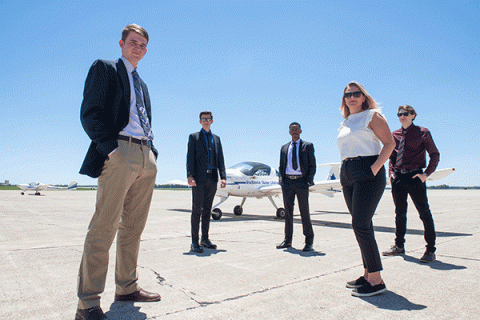 Four men and one woman stand on an open airport tarmac. A white airplane with blue lettering that says Indiana State University is visible behind them. The sky is blue and the day is bright and sunny. Another white plane is visible far in the distance on the left of the image.