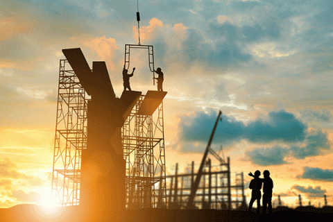 The frame and scaffolding of a building under construction is backlit by the rising sun. Two workers at the very top lift the arms to steady a section of frame being lowered to them by a construction crane (not visible), while two workers on the ground talk and one motions toward the building frame with his hand. All workers are in shadow but can be seen to wear hardhats.