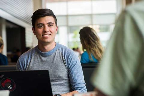 A man smiles while sitting at a table with a laptop