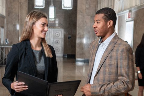 A white female student smiling in professional attire and holding a folder talking to a Black student in professional attire in the hallway of a building.