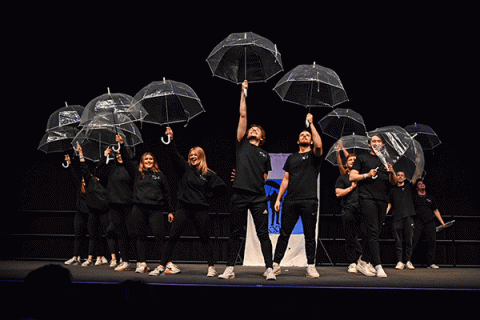 A group of male and female students dressed in black stand illuminated on a dark stage as they raise clear plastic umbrellas above their heads. A blue-and-white banner is visible behind them. One man on the right side holds his umbrella at a left angle to the side and not above his head like the others.