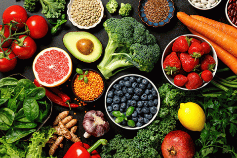 A colorful assortment of fruits and vegetables on a table. Visible items include: red tomatoes on the vine; an avocado cut in half; a pink grapefruit cut in half; green, leafy spinach; lettuce; brown parsnips; bowls of blueberries and strawberries; broccoli; a dish of white beans; a dish of seeds; lemons; a red pepper; a purple and white mushroom; a pomegranate; and some carrots, among other foods.