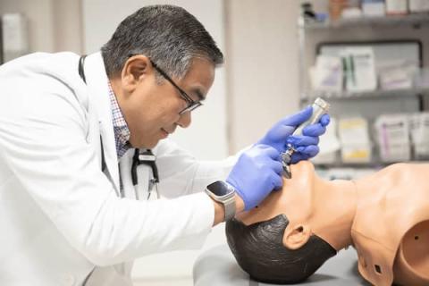 Middle aged male with white coat using a medical instrument to look down the throat of a medical practice mannequin.