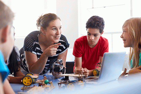 A young white female teacher with dark brown hair pulled into a ponytail smiles and points as she speaks with a group of children. She holds in her right hand a small toy vehicle with wheels and wires on it. On the table in front of the children are other, similar toys with yellow tires. To her left, a young boy with black, curly hair looks down at one of the toys as he listens. A grey laptop is open in front of him. Next to him, a young white girl with blonde hair smiles widely as she listens.