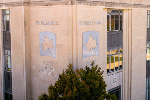 Aerial image of the corner of a large square building with a large metallic indiana state university sycamore leaf logo and the words “federal hall” above the leaf and “scott college of business” below the leaf with green trees in the foreground and on the side of the building.
