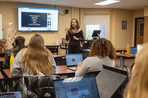 Female professor with dark hair and glasses standing in front of a classroom with back to camera. In the background is a human skeleton on a stand and a video board with medical information.