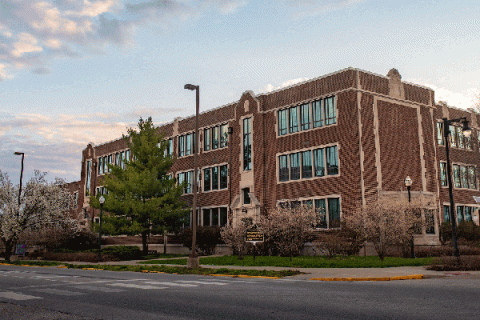 Exterior of a brick, three story building with large windows and trees in front with a blue sky and clouds and the intersection of two roads in front of the building