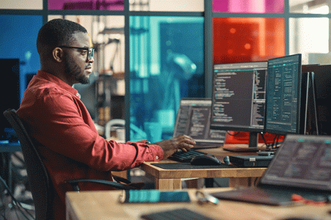 A Black male student sits at a desk in a computer lab. Two computer monitors are on a brown desk in front of him. The student has short black hair and matching facial hair. He wears black glasses and a red long-sleeved dress shirt. He sits in a black desk chair, typing on a black keyboard. Brown desks with computer monitors are also visible in the lab. Blue, red, purple, pink, and clear-colored glass windows are in the background. 