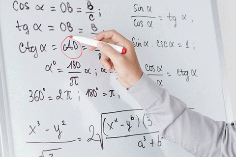 A close-up picture of a whiteboard with multiple equations written on the board in black lettering. A white individual’s hand is holding a red dry erase marker to the whiteboard, circling an equation. The person is wearing a long-sleeved white dress shirt.   