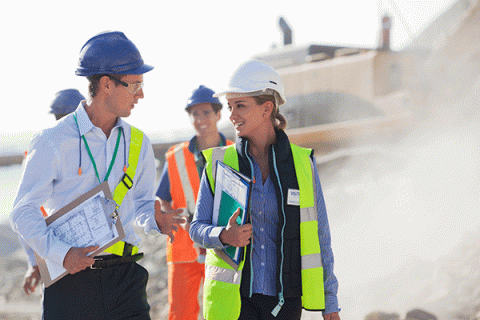 Two individuals are walking at a construction site. On the left is a white male with short brown hair. He holds a clipboard with a white paper document in his right hand. On the right is a white female with blond hair pulled back into a ponytail. The two individuals are looking at each other as they walk. Behind them is a white male wearing a blue hard hat and an orange full-body safety outfit with a blue hard hat. A bulldozer is in the background. 