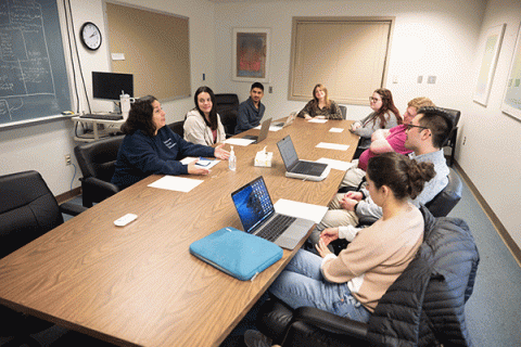 In a conference room, there are eight individuals of mixed races sitting at a table. They are wearing a variety of grey, white, blue, and pink clothing. Laptops are on the brown conference table. 