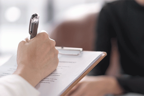 Closeup image of a right hand holding a pen and writing on a form with a clipboard, with a person sitting on a couch out of focus in the background. 