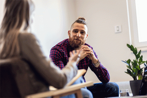 A white male with a beard and hair in a bun and facing the camera listens as a counselor in the foreground with her back to the camera and slightly out of focus speaks. 