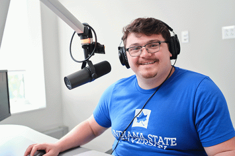 A white male student with short brown hair and glasses wearing a blue t-shirt with “Indiana State University” in white lettering. He looks and looking directly at the camera while wearing headphones and sitting in front of a radio microphone. 