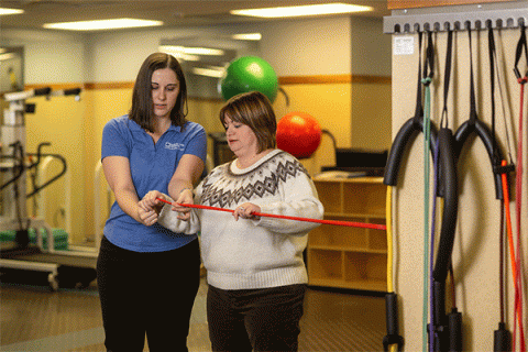 On the left is a white female student with shoulder-length brown hair wearing a blue shirt and black pants. She works with a female client on her right in a physical therapy clinic. The client is a white woman with shoulder-length brown hair wearing a white and brown sweater and black pants. They’re holding a red elastic physical therapy band and other equipment is visible in the background. 
