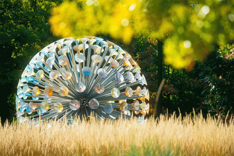 Photo of a large, circular structure made up of mall colorful disks suspended by metal posts installed in a round core, with brown native grass in the foreground and green foliage from a tree in the top right corner. 