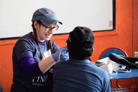 A white female student with a denim hat and glasses wearing blue surgical gloves holds an instrument towards a young patient with his back turned to the camera in a rural South American clinic with orange walls and a whiteboard in the background. 