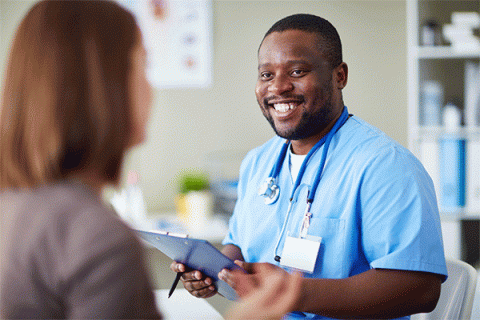 Black male with short hair, beard and mustache wearing blue nursing scrubs and a nametag and holding a clipboard smiles and talks to a female patient with her back to the camera in a medical office setting.   