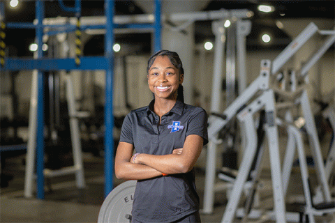 Black female wearing black polo with the Sycamores logo standing with arms crossed and smiling at the camera with exercise equipment in the background. 