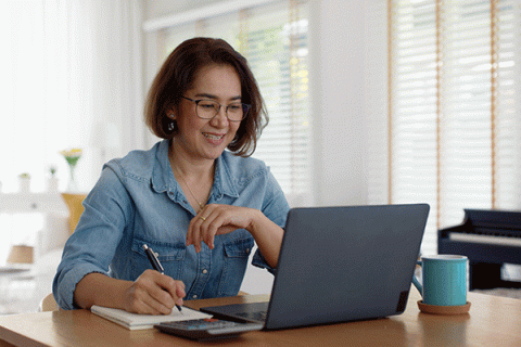 Middle-aged female of Asian descent with short brown hair, glasses, and wearing a denim shirt while sitting at a table with a laptop and writing on a notepad with her right hand. A calculator is visible on the table.  