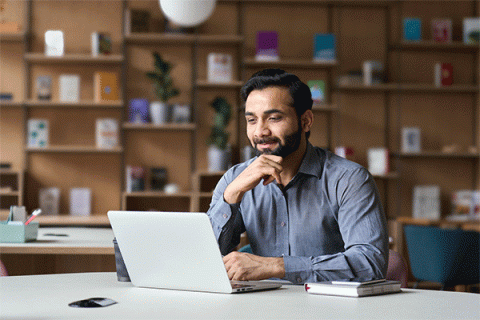 Male with dark hair, beard and mustache in a dress shirt sitting at a large table and looking down at a laptop screen.