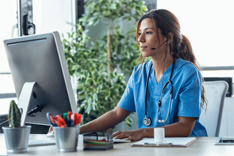 Female with long brown hair wearing blue nursing scrubs looking at a desktop computer screen while sitting at a table.  