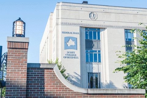 Large, multi-story sandstone building with metal lettering reading “Federal Hall” and “Scott College of Business” with a metallic Sycamores logo, with blue sky and a brick structure visible in the foreground. 