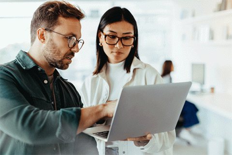 A man with brown hair, a beard and mustache, and glasses, wearing a black shirt, points at a laptop while a woman with straight, shoulder-length, dark brown hair and glasses, wearing a white turtleneck and white jacket, holds the laptop. They are in a bright office setting.  