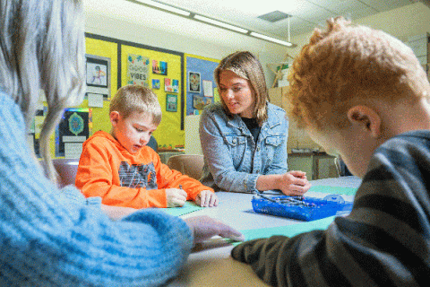 A blonde teaching student with short hair and wearing a blue jean jacket is seated at a table in a classroom. She watches as a young blond boy in an orange sweatshirt seated next to her creates a drawing on green construction paper. In the foreground, another young boy with short red hair works on a drawing, while another female teaching student with long blonde hair and wearing a blue sweater holds the paper in place for him. 