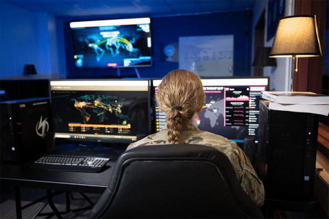 Blond female wearing fatigues seated with back turned to the camera working on a computer with monitors depicting global threats on various maps.