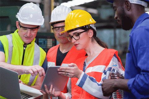 Diverse group of workers wearing hard hats and safety vests look at data on a tablet in a factory setting. 