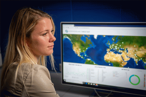 Blonde female student seated in front of a computer monitor and looking to the right with a map showing cybercrime activity on a screen. 