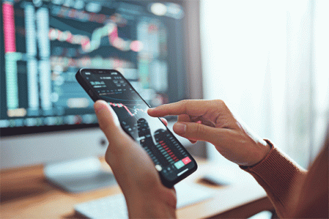 Detail image of person holding a phone with financial information on the screen with more information on a computer screen in the background. 