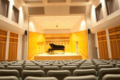 An empty recital hall with a piano in the center of a stage in the background, with rows of seats in the foreground.