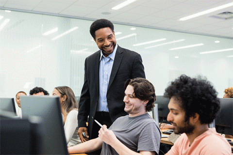 Smiling mature adult male university professor checks in with two male students in a computer lab setting.