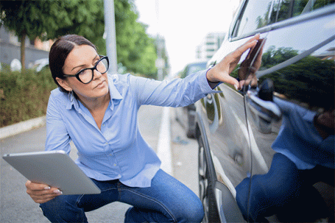 Female middle-aged woman with a tablet in her hand kneeling down and inspecting minor damage on the door of a vehicle. 