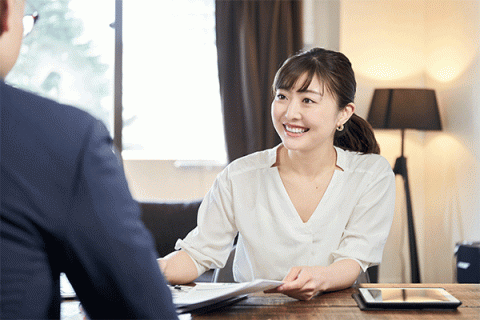 Middle-aged woman of Asian descent sitting at a table and smiling at a client across the table with back to the camera in an office setting.