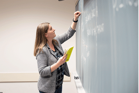 Female professor in a classroom using her left hand to write on a chalkboard in front of a class. 