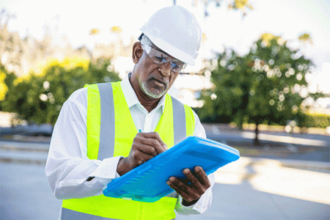 A senior black male construction site manager writing on a blue tablet while wearing a hard hat, safety goggles and a yellow construction vest while inspecting a building project