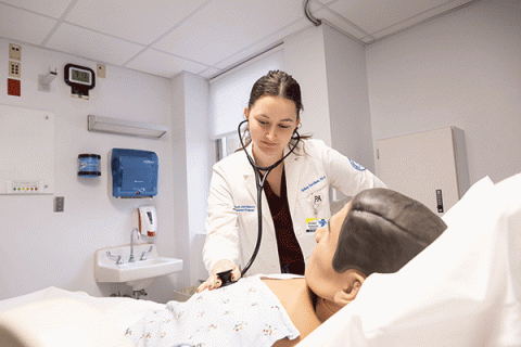Female student in a white lab coat uses a stethoscope to check vitals on a human simulation dummy in a hospital bed in a white lab setting. 