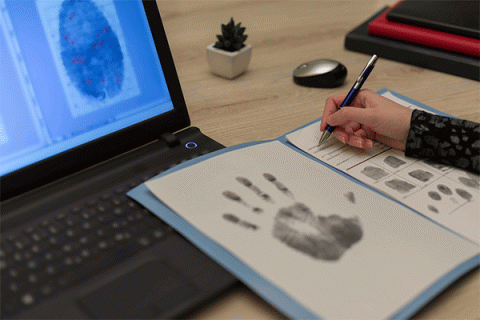 Detailed image of the hand of a student writing on a form containing a full handprint and fingerprints while viewing a fingerprint on a laptop.