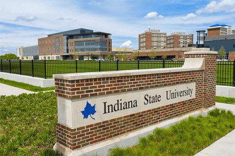 A large brick building with large windows is visible in the background of the image on the left, with three campus towers in the distance on the far right of the frame. In the foreground is a brick campus entryway surrounded by green grass, with a blue Sycamore leaf logo and “Indiana State University” on the sign. 