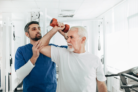 Senior man with white hair lifting a dumbbell, with assistance from an occupational therapist in a medical setting. 