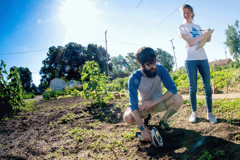 A MALE STUDENT KNEELS ON THE GROUND AND CHECKS THE SOIL WITH AN INSTRUMENT WHILE A FEMALE STUDENT STANDING NEARBY TAKES NOTES IN A GARDEN SETTING. 