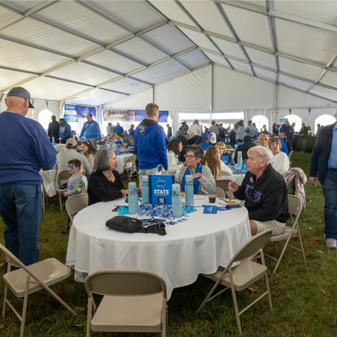 A large group of Indiana State University alumni and friends gather under a large tent for tailgating, sitting around tables with drinks and snacks, wearing ISU gear.