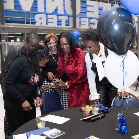 A group of five women, dressed formally, gather around a table looking at a smartphone during a Black Alumni Reception at Indiana State University. Blue and white balloons decorate the table.