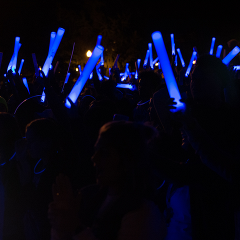 Attendees at the Blue Light Party at Indiana State University hold up glowing blue sticks in a darkened outdoor setting, creating a vibrant, energetic atmosphere.