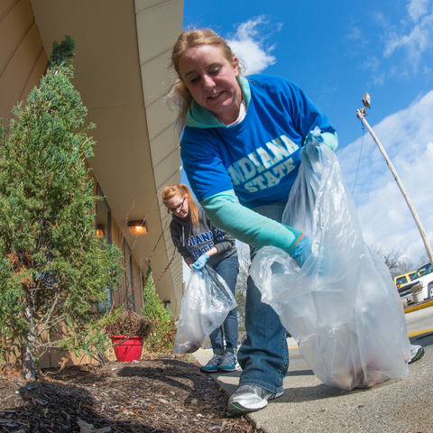 wo volunteers in Indiana State University shirts clean up outside a building, collecting trash in large plastic bags on a sunny day.