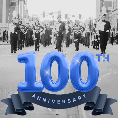 A black and white photo of a marching band parading down a street, led by a drum major. In the foreground, a large blue balloon-style text reads '100th Anniversary' with a blue ribbon banner below it.